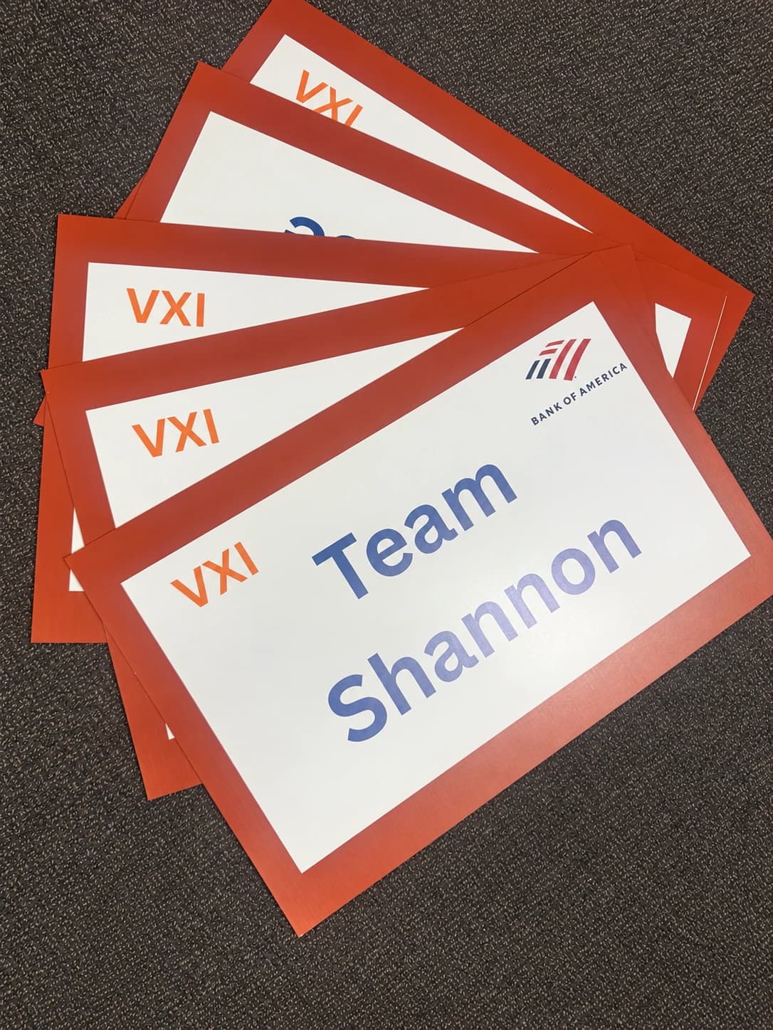 Overlapping red-bordered foam-board signs with VXI and Bank of America branding; the front sign reads Team Shannon.